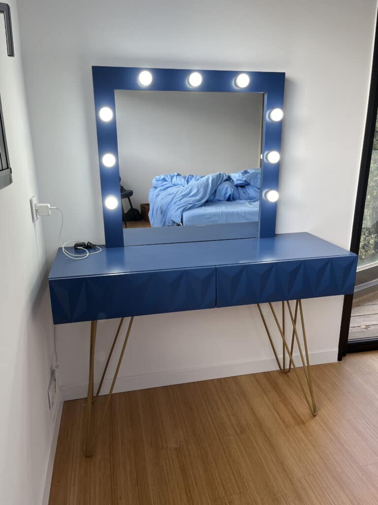 Coiffeuse Miroir Hollywood Bleu et Pieds Dorés Moderne Coiffeuse bleu marine moderne avec miroir éclairé et tiroirs géométriques. Pieds épingles dorés, chargeur visible sur le plateau.
